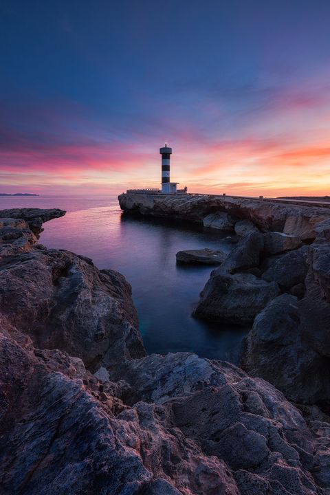 Lighthouse on rocky coastline at sunset with vivid purple, pink, and orange sky reflecting on calm sea.