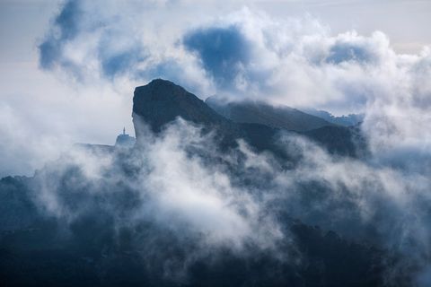 Dramatic mountain peak surrounded by swirling clouds with a distant lighthouse visible on a cliff, Mallorca, Spain.