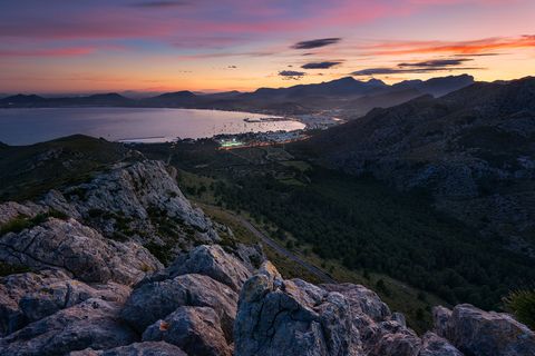 Rocky mountain viewpoint overlooking a coastal town and bay at sunset, with illuminated buildings and dramatic sky in Mallorca, Spain.