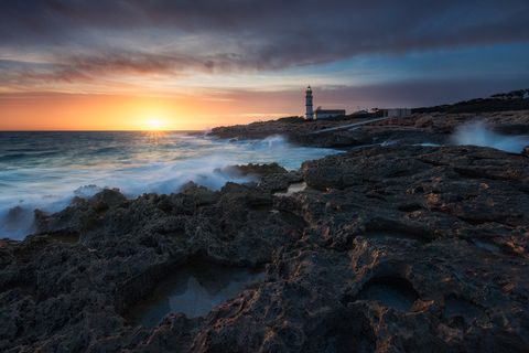 Rocky coastline with waves crashing against the shore at sunset, a lighthouse stands in the background under a dramatic sky.