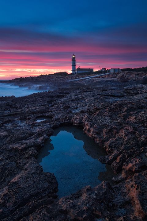 Rocky coastline with a tidal pool in the foreground, a lighthouse in the distance, and a vibrant colorful sunset sky.