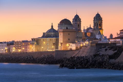 Sunset view of Cádiz Cathedral with its domes and towers, city lights, and waterfront in Cádiz, Spain.
