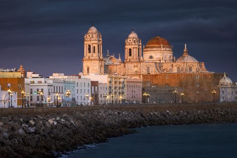 View of the Cádiz Cathedral and colorful buildings along the waterfront in Cádiz, Spain, under dramatic dark clouds at sunset.