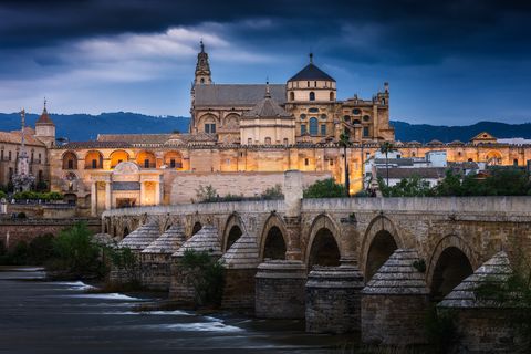 Historic Roman bridge and the illuminated Mezquita-Catedral of Córdoba at dusk, under a dramatic cloudy sky.