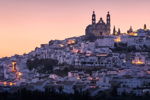 Whitewashed houses in Olvera with a large church and twin bell towers above, illuminated by warm lights at sunset in Spain.
