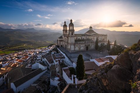 Sunset view of a hilltop church with twin towers overlooking white-washed houses in a Spanish village, surrounded by rolling green hills and mountains.