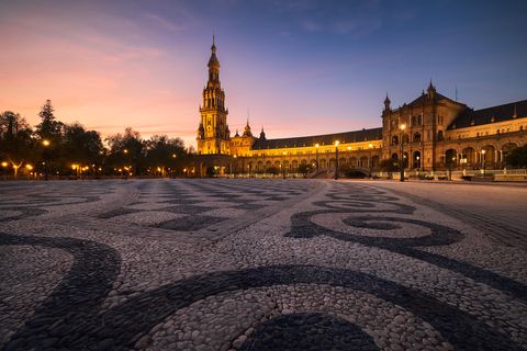 Plaza de España in Seville at sunset, featuring ornate tilework on the ground and the illuminated central tower and building.