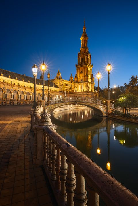Plaza de España in Seville, Spain, illuminated at dusk with glowing street lamps, a decorative arched bridge over a canal, and the reflection of the tower in the water.