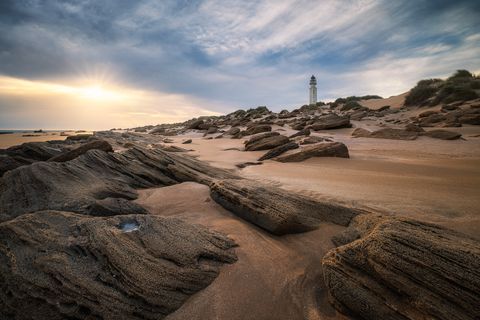 Rocky sandy beach with large stones, sunset sky, and the white Trafalgar lighthouse surrounded by dunes and sparse vegetation, Andalusua, Spain.