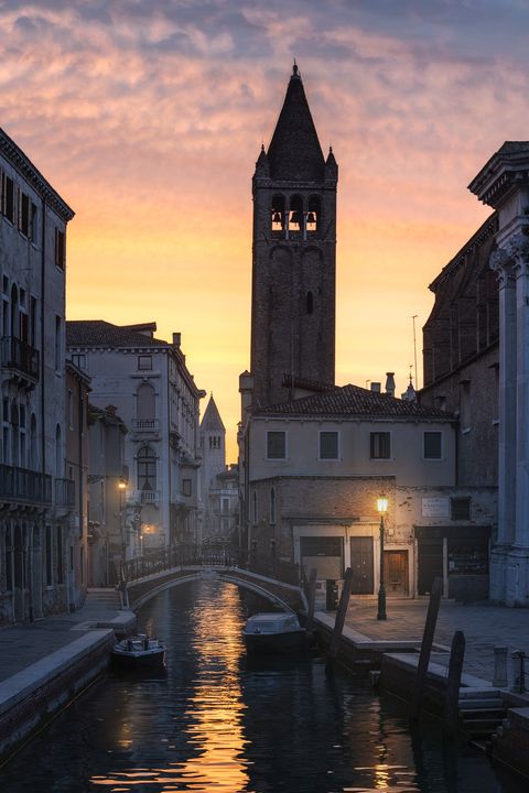 Venetian canal at sunrise with boats, a stone bridge, historic buildings, and a tall bell tower silhouetted against a vibrant sky.