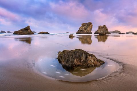 Large sea stacks and rocks reflected in wet sand at Bandon Beach during a colorful sunrise with pink and purple clouds in the sky.