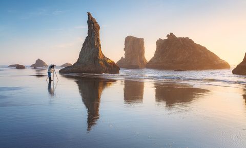 Sunrise at Bandon Beach, Oregon with sea stacks reflected in wet sand and a lone photographer taking pictures with a tripod.