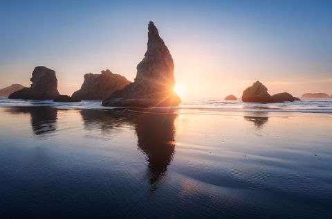 Sea stacks and rocky formations at Bandon Beach at sunset, with sunlight reflecting on wet sand and calm ocean waves.
