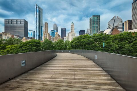 Wooden pedestrian bridge curving through green trees with downtown Chicago skyscrapers and cloudy sky in the background.