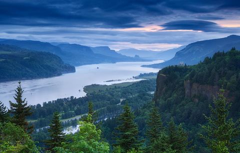 Scenic view of the Columbia River Gorge with lush green forests, dramatic cliffs, and cloudy sky at sunrise or sunset.