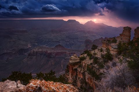 Sunrise over the Grand Canyon with dramatic clouds, layered rock formations, and rugged cliffs in the foreground.
