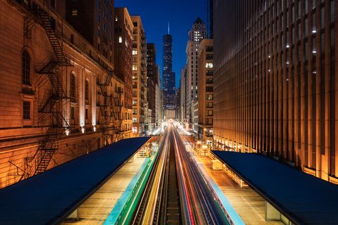 Night view of Adams & Wabash train station in Chicago with light trails from trains, surrounded by tall buildings and a skyscraper in the background.