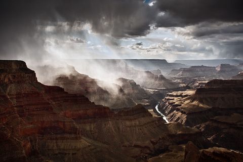 Dramatic view of the Grand Canyon with sunlight streaming through dark storm clouds and mist over red rock formations and a river below.