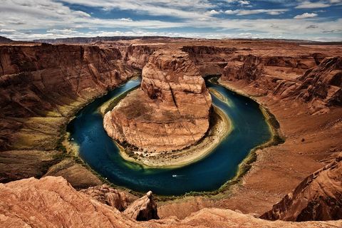 Aerial view of Horseshoe Bend in Arizona with the Colorado River curving around a large red rock formation under a partly cloudy sky.