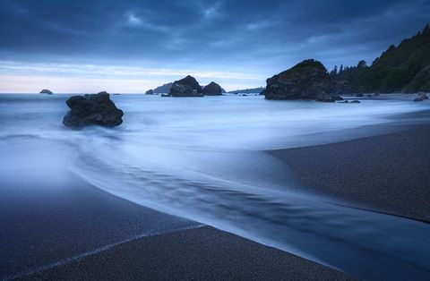 Rocky coastline at dusk with dark sand, flowing water, sea stacks, and a dramatic cloudy sky.