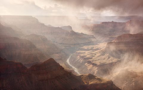 A dramatic view of the Grand Canyon with mist and sunlight filtering through clouds, revealing layered rock formations and a winding river below.