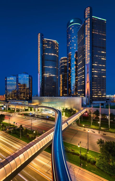 Nighttime cityscape of Detroit with illuminated skyscrapers, including the Renaissance Center, and a curving elevated people mover track in the foreground.