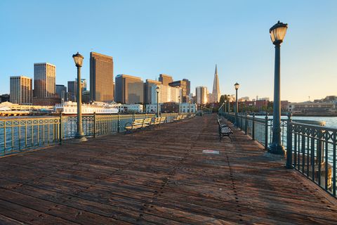 Wooden pier with benches and street lamps overlooking the San Francisco skyline, including the Transamerica Pyramid, at sunset.