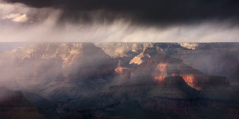 Dramatic storm clouds and mist hover over the Grand Canyon, with sunlight illuminating the rocky canyon walls and highlighting rich red and orange hues.