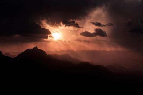 Dramatic sunset over silhouetted mountains with dark clouds and rays of sunlight breaking through.