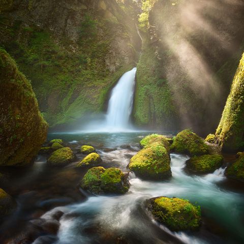 Wahclella waterfall cascading into a clear pool surrounded by moss-covered rocks and lush green cliffs, with sunlight streaming through mist.