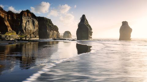 Meeresfelsen und zerklüftete Klippen spiegeln sich im nassen Sand bei Sonnenuntergang am Three Sisters Beach auf der Nordinsel Neuseelands.